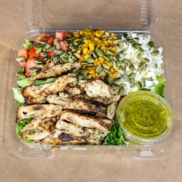 Grilled chicken salad with corn, tomatoes, pumpkin seeds, and green dressing in a clear deli container.